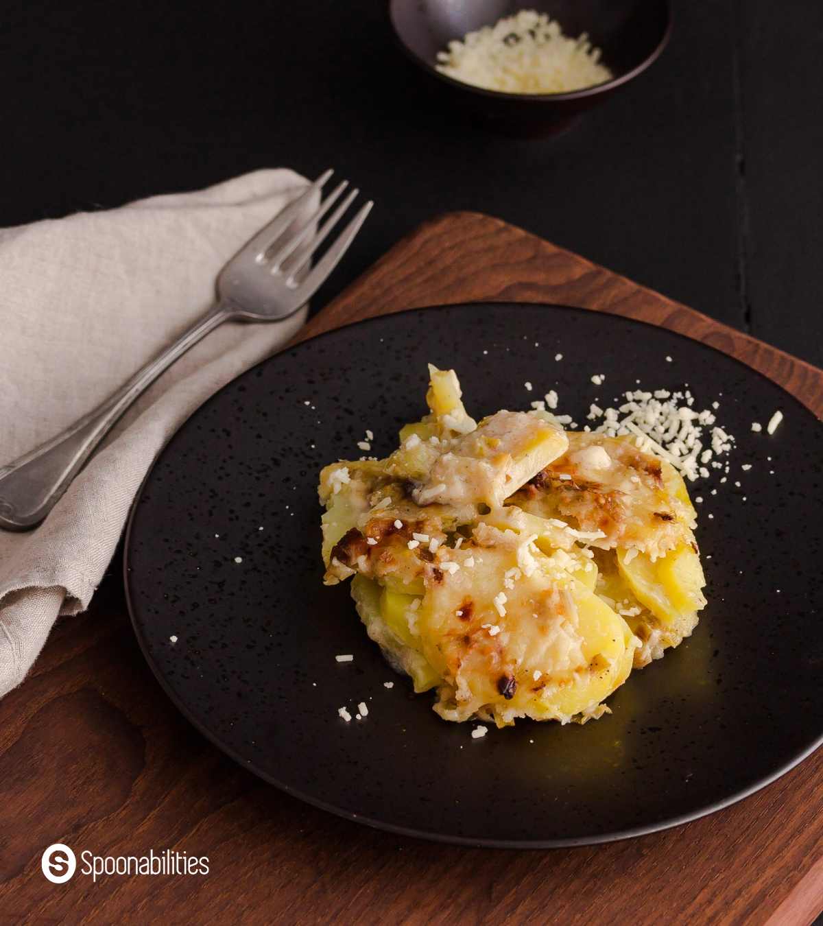 Leek and potato gratin on a black plate with a fork on the side and some cheese for topping on the side