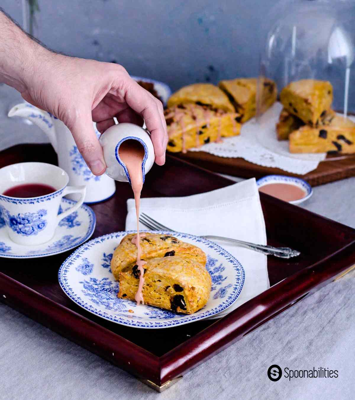 Pouring blood orange maple syrup glaze on pumpkin scones on a white and blue plate with tea on the side
