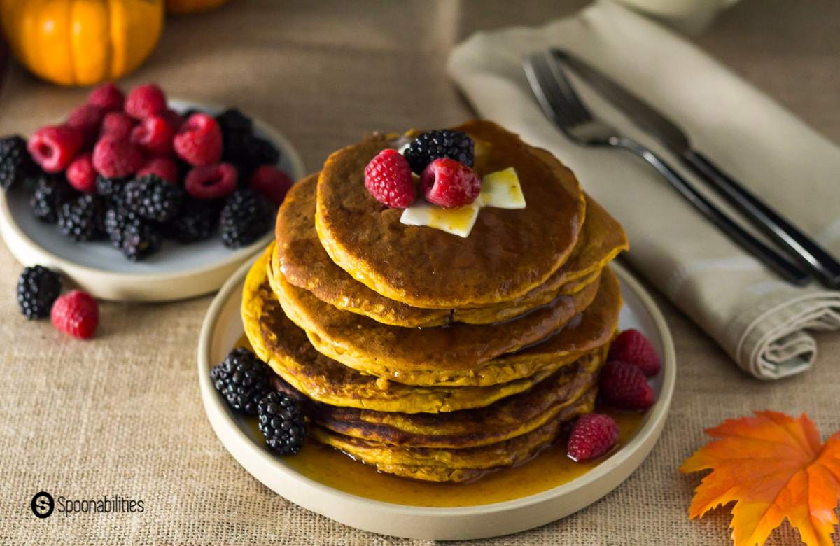 Pumpkin spice pancakes stack on a round white plate with a maple leaf in the foreground and mixed berries in a plate in the background