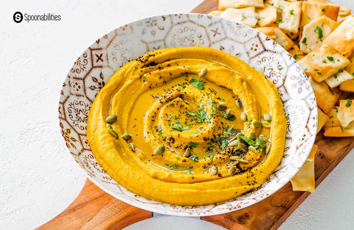 Pumpkin hummus fall dip on a white plate with gold accents on wooden board with pita chips