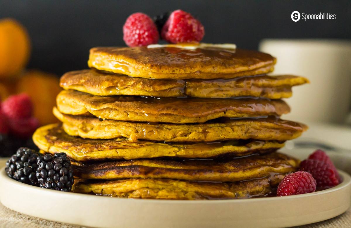 Seven spiced pumpkin pancakes stacked on top each other on a white round plate with syrup and berries