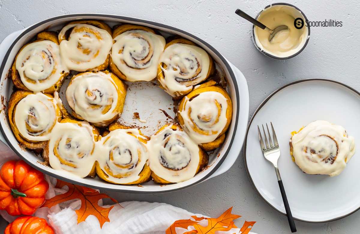 a pumpkin cinnamon roll in a white plate taken from a baking dish full of cinnamon rolls with icing