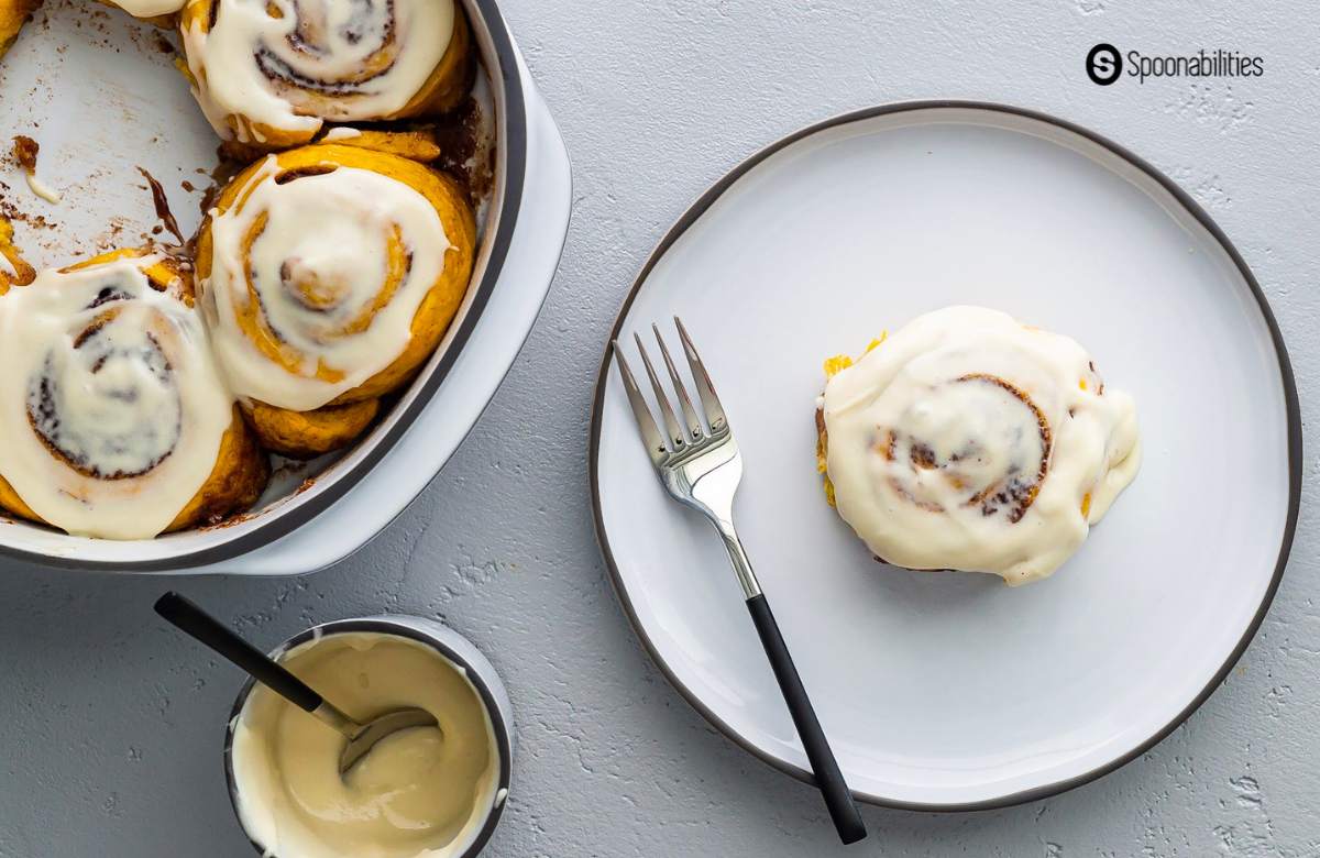 pumpkin cinnamon roll on a plate with cream cheese icing and more icing in cup on the side