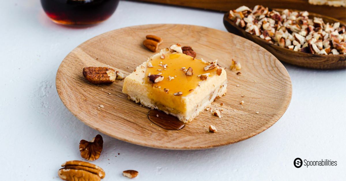 Cheesecake bar on a wooden plate with maple syrup and crushed pecan toppings and small bowl of crushed pecan on the side