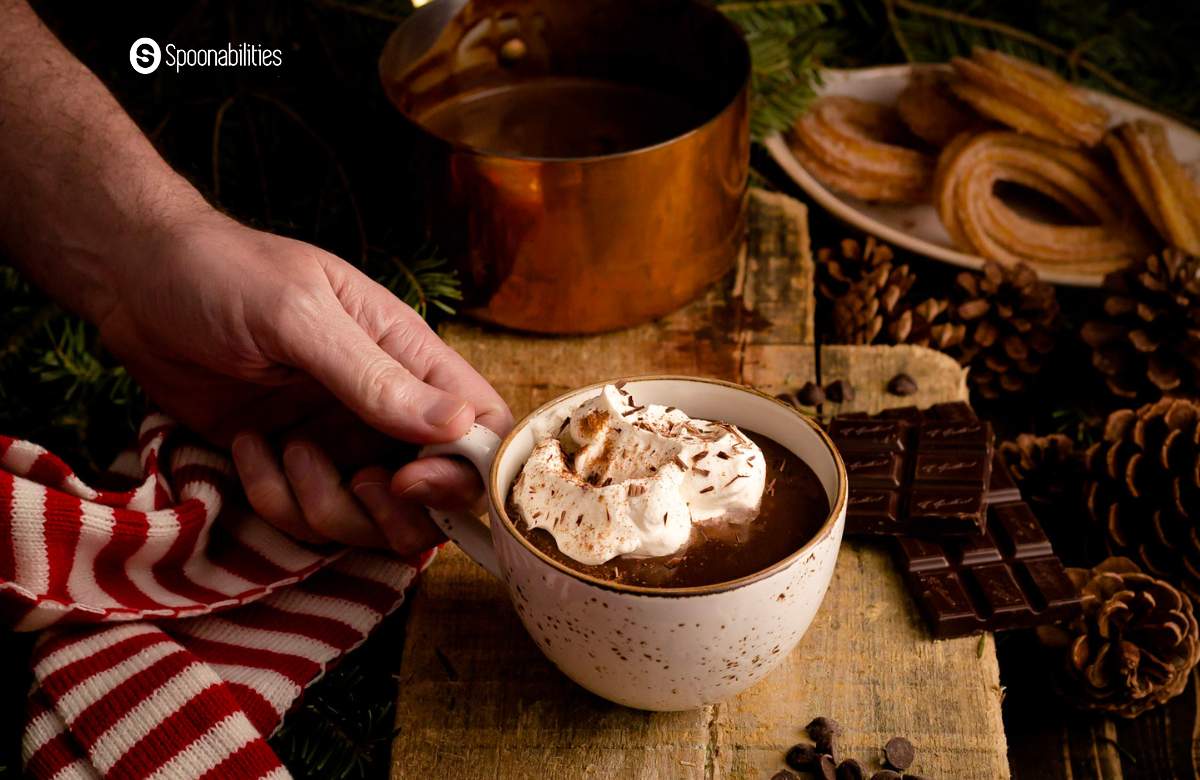 A hand placing a cup of Spanish Hot Chocolate with whipped cream on a wooden plank with chocolate bars and chips scattered around it